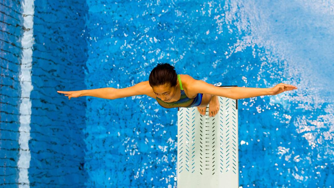 Junge Sportlerin steht auf einem Sprungbrett, bereit zum Sprung in einem blauen Schwimmbecken.