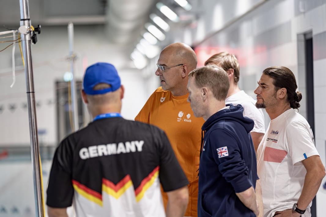 Fünf Trainer aus verschiedenen Nationen beobachten ihre Athleten im Wasser.