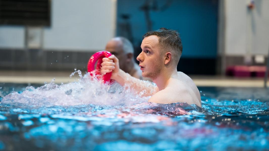 Mann im Wasser hält einen pinken Kegel und spritzt Wasser, im Hintergrund eine weitere Person.