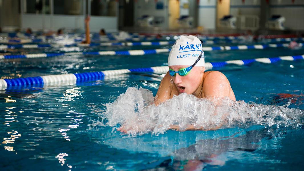 Schwimmerin in Aktion beim Brustschwimmen in einem Innenbecken, mit spritzendem Wasser und Schwimmbrille.