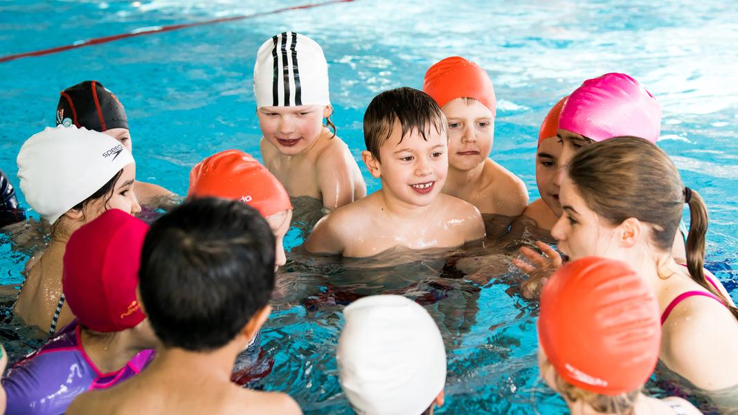 Gruppe von Kindern im Schwimmbecken, die sich um eine Trainerin versammeln und miteinander kommunizieren.