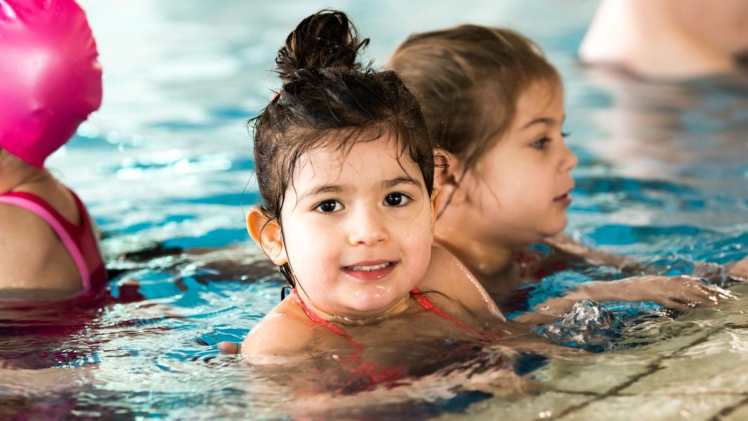 Lächelndes Mädchen mit nassen Haaren im Wasser, im Hintergrund ein weiteres Kind in der Schwimmhalle.