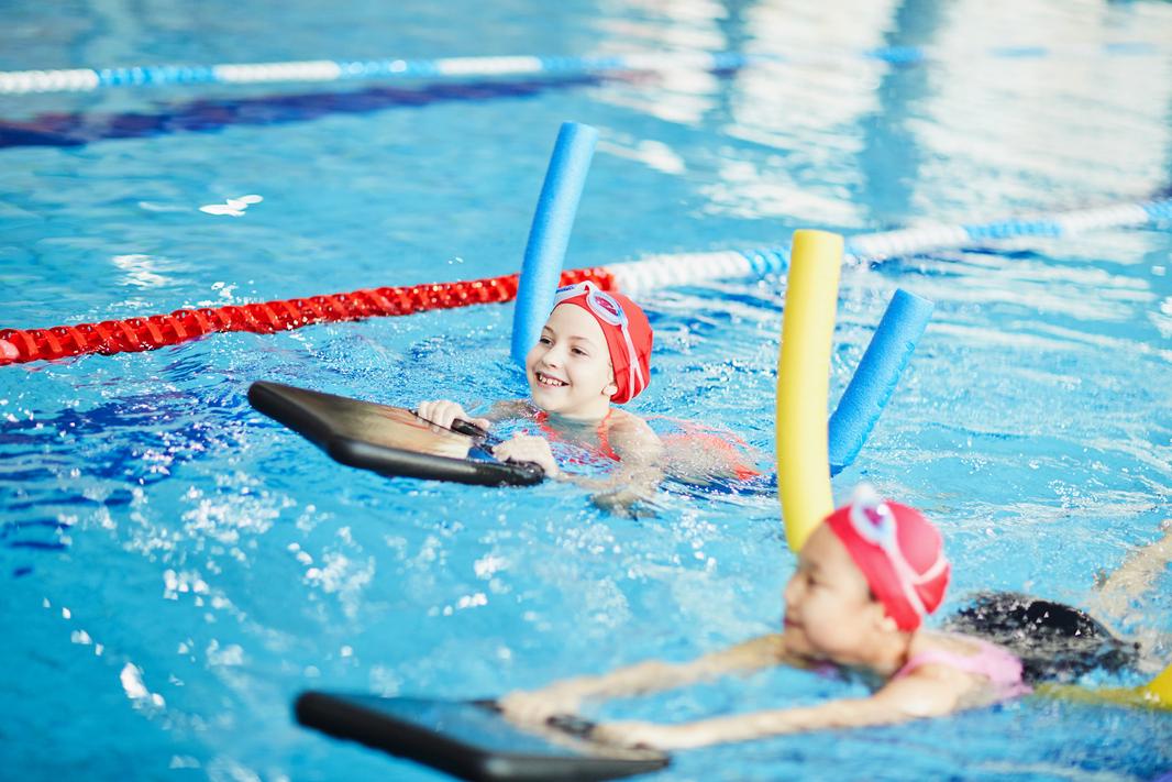 Zwei Kinder mit Schwimmnudeln schwimmen im Pool, eines lächelt und hält ein Schwimmbrett in der Hand.